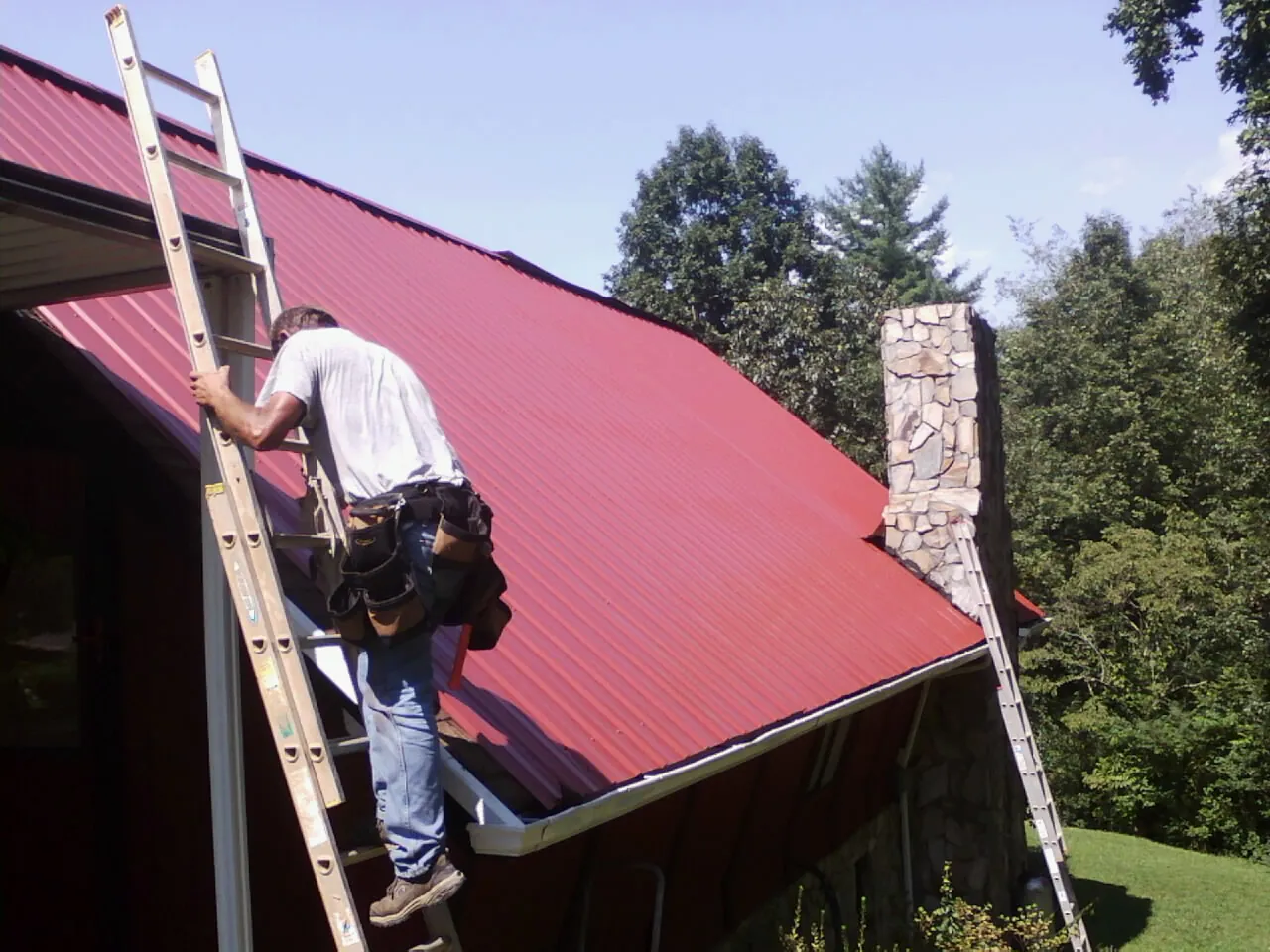 Roofer on a ladder beside a red metal roof and stone chimney — Soffit & Fascia Repair by Roofing Brothers in Haven